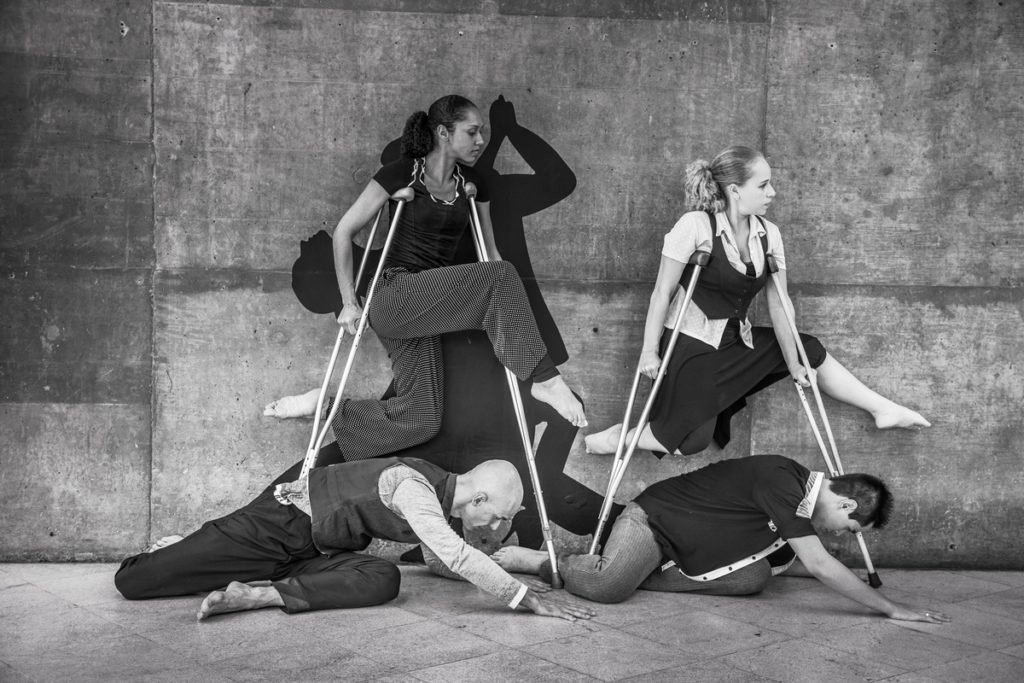 Descrição da imagem #PraCegoVer: A imagem preto e branco está no formato retangular, na horizontal. Nela, os atores da companhia de dança estão em uma performance. Duas mulheres parecem pairar no ar, apoiadas em muletas. No chão, dois homens parecem se rasteijar abaixo delas. Fim da descrição.