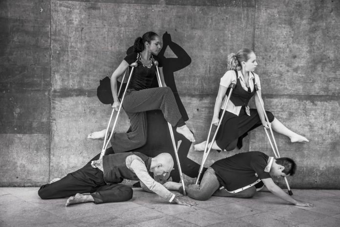 Descrição da imagem #PraCegoVer: A imagem preto e branco está no formato retangular, na horizontal. Nela, os atores da companhia de dança estão em uma performance. Duas mulheres parecem pairar no ar, apoiadas em muletas. No chão, dois homens parecem se rasteijar abaixo delas. Fim da descrição.