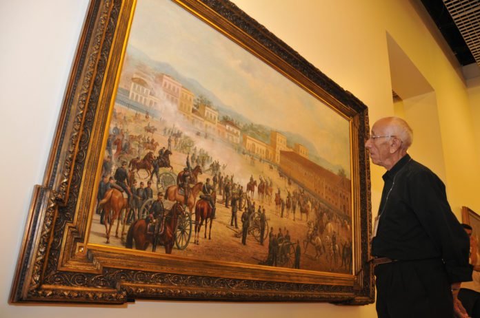 Descrição da imagem #PraCegoVer: A imagem está no formato retangular, na horizontal. Nela, um senhor observa um quadro em um museu. Fim da descrição.