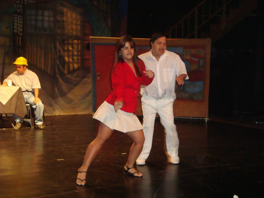 Descrição da imagem #PraCegoVer: A imagem está no formato retangular, na horizontal. Nela estão dois atores com síndrome de Down, que integram a companhia de teatro. Eles estão dançando, no centro do palco, durante uma apresentação. Fim da descrição.
