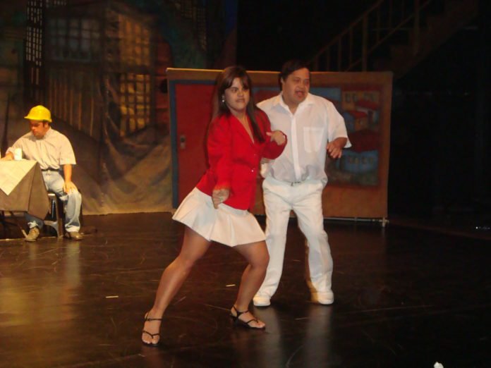 Descrição da imagem #PraCegoVer: A imagem está no formato retangular, na horizontal. Nela estão dois atores com síndrome de Down, que integram a companhia de teatro. Eles estão dançando, no centro do palco, durante uma apresentação. Fim da descrição.