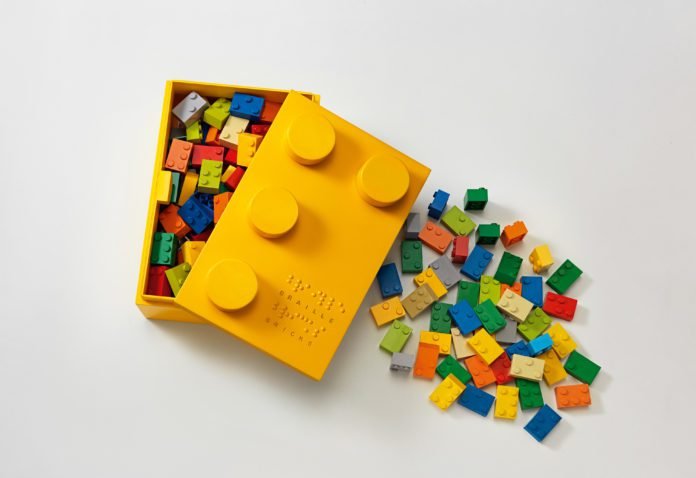Descrição da imagem #pracegover: Foto da caixa do Braille Bricks. Ela é amarela e está semiaberta, sendo possível ver os blocos de montar coloridos em seu interior. Do lado direito da caixa, algumas outras peças estão espalhadas. Fim da descrição.