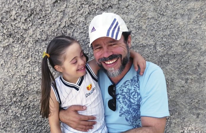 Descrição da imagem #PraTodosVerem: A imagem está no formato retangular, na horizontal. Nela estão a pequena Ana Luíza e seu pai, o artista Elitan. Eles estão abraçados e ambos sorriem. Fim da descrição.
