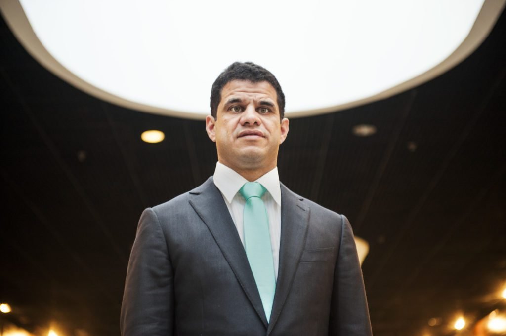 Descrição da imagem #PraCegoVer: Imagem no formato retangular, na horizontal. Foto de close de Mizael Conrado. Ele usa um terno preto, camisa branca e gravata verde. Fim da descrição.