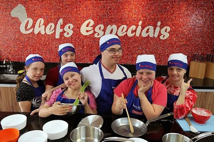Descrição da imagem #PraCegoVer: A imagem está no formato retangular, na horizontal. Nela, seis jovens com deficiência estão na cozinha do instituto Chefs Especiais. Eles estão usando uniforme de cozinheiro e alguns seguram utensílios de cozinha. Todos estão sorrindo. Fim da descrição.