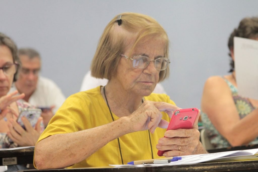 Descrição da imagem #PraCegoVer: A imagem está no formato retangular, na horizontal. Nela, uma idosa está sentada em uma sala de aula, manuseando um telefone celular. Fim da descrição.