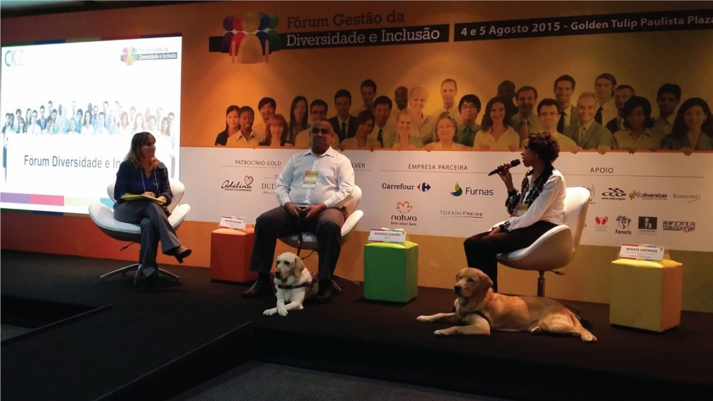 Descrição da imagem #PraCegoVer: A imagem está no formato retangular, na horizontal. Nela estão três pessoas sentadas em poltronas brancas, que participam como palestrantes do evento. Entre eles estão duas mulheres e um homem. A mulher que está sentada à esquerda está sozinha. Já o homem que está sentado no meio e a outra mulher, que está do lado direito, estão acompanhados de seus cães-guias. Ao fundo, está um painel com os logotipos de patrocinadores e informações do evento, que discutiu a diversidade e a inclusão de pessoas com deficiência no mercado de trabalho. Fim da descrição.