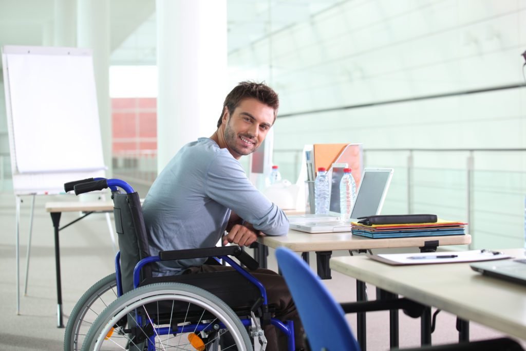 Descrição da imagem #PraCegoVer: A imagem está no formato retangular, na horizontal. Nela, um jovem rapaz está sentado em sua cadeira de rodas. Ele está em seu ambiente de trabalho, um escritório. Ele olha para o lado e sorri. Fim da descrição.