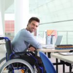 Descrição da imagem #PraCegoVer: A imagem está no formato  retangular, na horizontal. Nela, um jovem rapaz está sentado em sua cadeira de rodas. Ele está em seu ambiente de trabalho, um escritório. Ele olha para o lado e sorri. Fim da descrição.