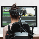 Descrição da imagem #PraCegoVer: A imagem está no formato  retangular, na horizontal. Nela é possível ver, uma mulher, sentada de costas em uma cadeira de rodas. Ela está sentada em frente a uma tela do computador. Fim da descrição.
