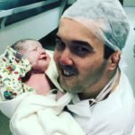 Descrição da imagem #PraCegoVer: Imagem no formato retangular, na horizontal. Leonardo Reis segura sua filha, Luísa, no colo. Luísa acabou de nascer e está embrulhada em uma mantinha amarela, com desenhos de palhaços. Ela está com os olhinhos fechados. Leonardo está com um avental e touca, itens obrigatórios para estar em um centro cirúrgico. Ele está sorrindo. Fim da descrição.