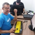 Descrição da imagem #PraCegoVer: Imagem no formato retangular, na horizontal. Um homem está em um escritório. Ele tem baixa visão e usa um computador com teclado adaptado. Além do monitor e do teclado do computador, em cima da mesa tem uma lupa. O homem é moreno, tem cabelos curtos e grisalhos. Ele usa uma camiseta na cor azul e usa óculos. Fim da descrição.