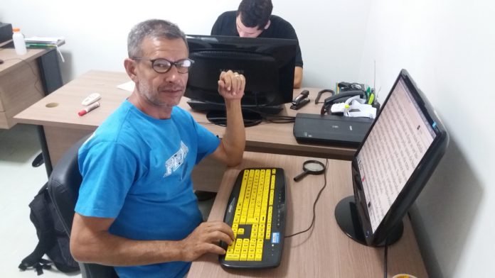Descrição da imagem #PraCegoVer: Imagem no formato retangular, na horizontal. Um homem está em um escritório. Ele tem baixa visão e usa um computador com teclado adaptado. Além do monitor e do teclado do computador, em cima da mesa tem uma lupa. O homem é moreno, tem cabelos curtos e grisalhos. Ele usa uma camiseta na cor azul e usa óculos. Fim da descrição.