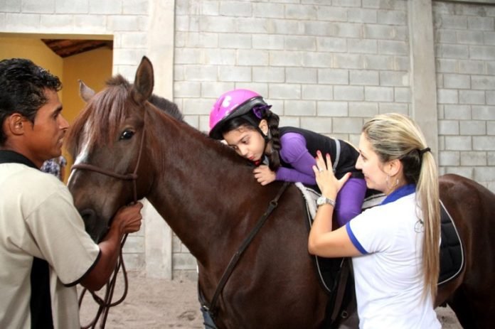 Descrição da imagem #PraCegoVer: Imagem no formato retangular, na horizontal. Nela, uma menina, paciente da equoterapia, está deitada em cima de um cavalo marrom. Um domador segura as rédeas do cavalo, enquanto uma equoterapeuta segura a menina durante o movimento. Fim da descrição.