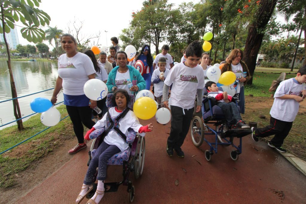 Descrição da imagem #PraCegoVer: A imagem está no formato retangular, na horizontal. Pais e filhos participam da segunda edição da caminhada inclusiva em um parque na cidade de Barueri. Todos usam uma camiseta branca e as crianças seguram bexigas coloridas, nas cores amarelo, branco e laranja. Na linha de frente, temos duas crianças cadeirantes, acompanhadas de seus pais. Ao fundo é possível ver algumas árvores. Fim da descrição.