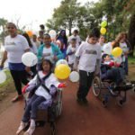 Descrição da imagem #PraCegoVer: A imagem está no formato retangular, na horizontal. Pais e filhos participam da segunda edição da caminhada inclusiva em um parque na cidade de Barueri. Todos usam uma camiseta branca e as crianças seguram bexigas coloridas, nas cores amarelo, branco e laranja. Na linha de frente, temos duas crianças cadeirantes, acompanhadas de seus pais. Ao fundo é possível ver algumas árvores. Fim da descrição.