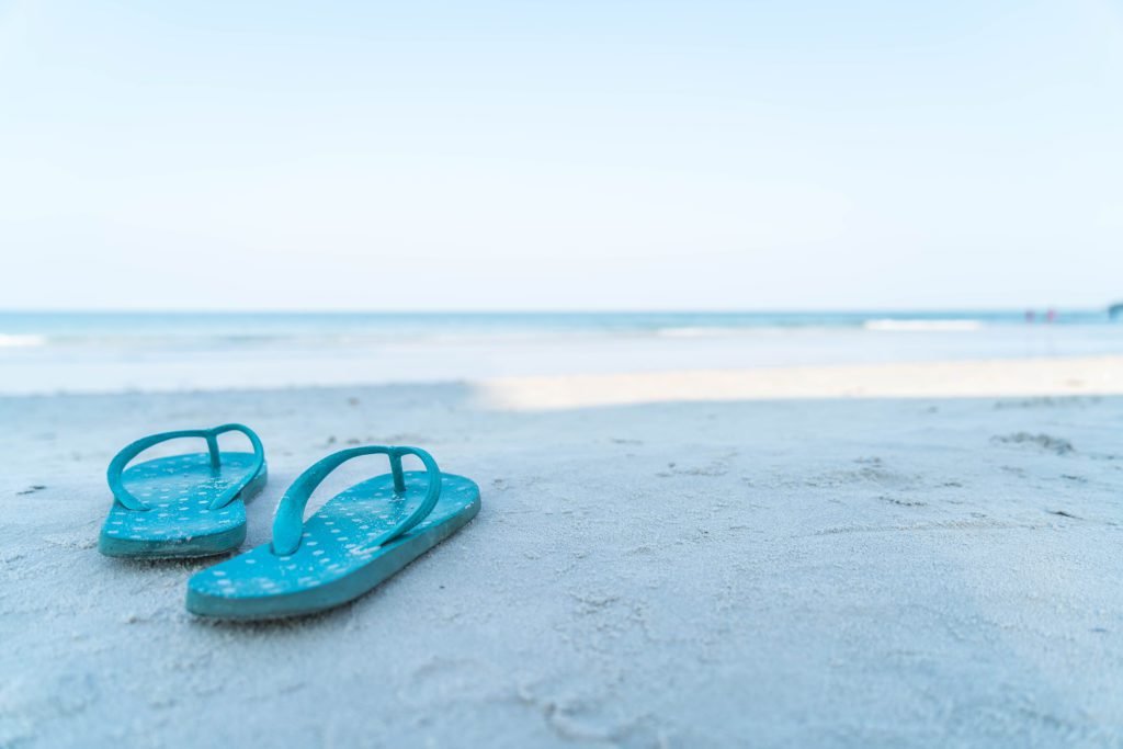 Descrição da imagem #PraCegoVer: Imagem no formato retangular, na horizontal. Nela, vemos uma praia. Em primeiro plano, do lado esquerdo da imagem, um par de chinelos na cor azul. Eles estão na faixa de areia da praia. Ao fundo, vemos o mar. Fim da descrição.