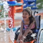 Descrição da imagem #PraCegoVer: Imagem no formato retangular, na horizontal. Nela, uma menina brinca com jatos d’água no parque aquático. Ela é morena tem cabelos ondulados e compridos. Ela é cadeirante e está sentada em uma cadeira de rodas. A menina está se divertindo muito. Fim da descrição.