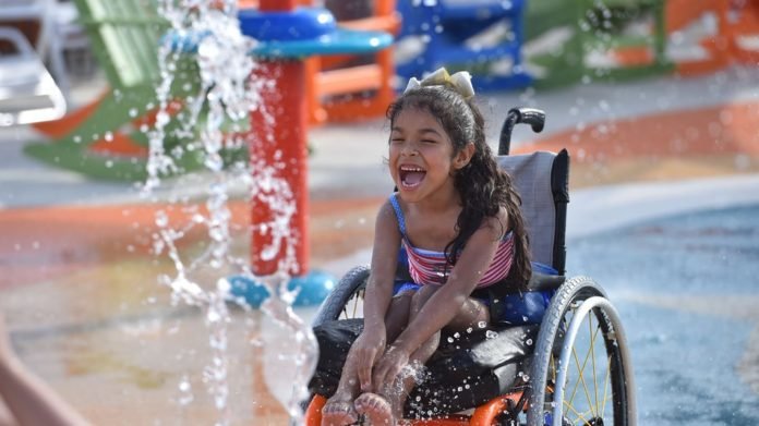 Descrição da imagem #PraCegoVer: Imagem no formato retangular, na horizontal. Nela, uma menina brinca com jatos d'água no parque aquático. Ela é morena tem cabelos ondulados e compridos. Ela é cadeirante e está sentada em uma cadeira de rodas. A menina está se divertindo muito. Fim da descrição.
