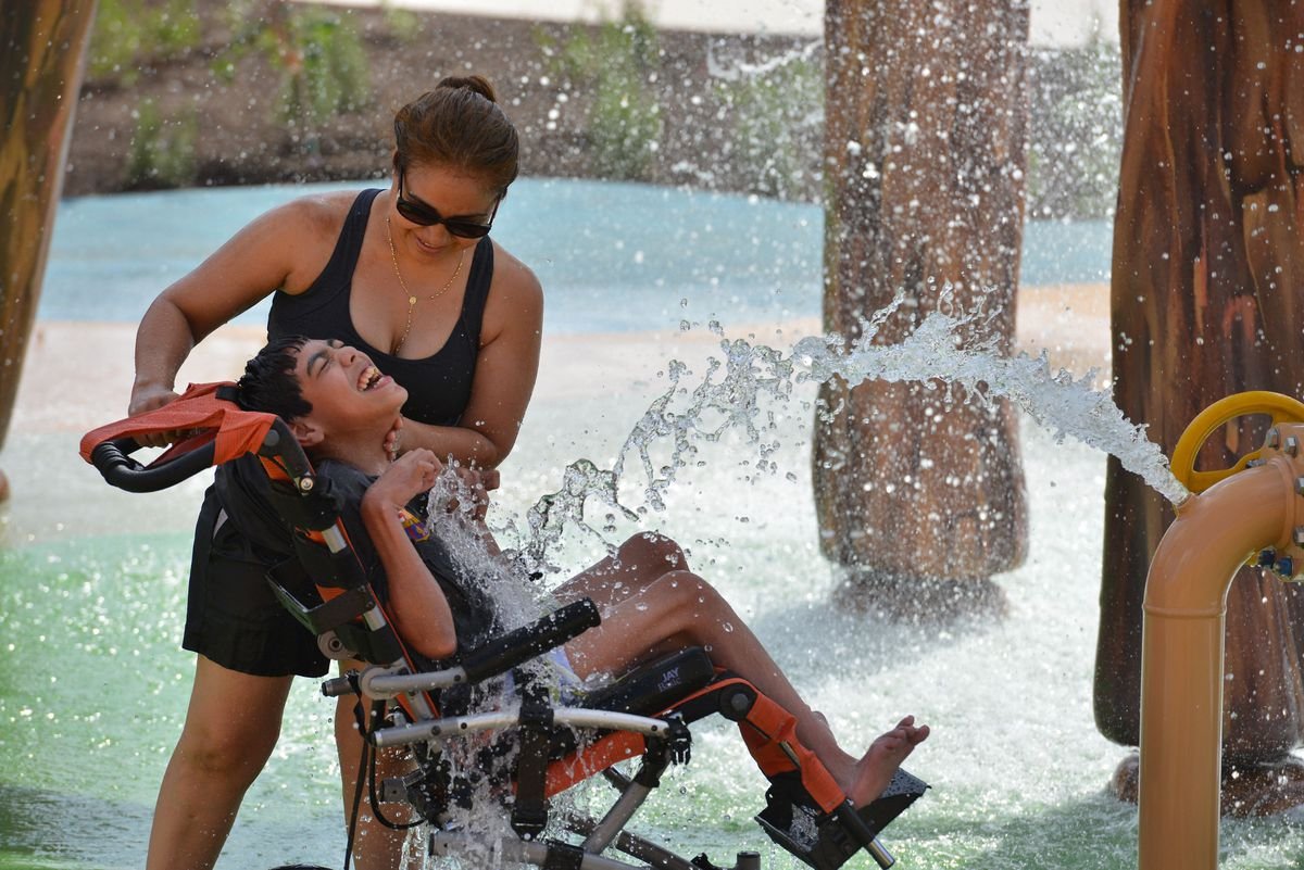 Descrição da imagem #PraCegoVer: Imagem no formato retangular, na horizontal. Nela, uma criança com paralisia cerebral se diverte ao lado de sua mãe. A criança brinca entre os jatos d'água do parque aquático. Fim da descrição.