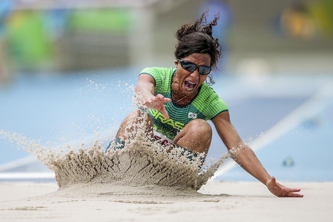 Descrição da imagem #PraCegoVer: Imagem no dormato retangular, na horizontal. A atleta Silvânia Costa, durante prova de salto em distância durante os Jogos Paralímpicos do Rio de Janeiro. O salto rendeu medalha de ouro para a atleta. Ela está exatamente no momento em que toca a areia durante seu salto. Ela tem deficiência visual e está com os olhos vendados, por uma regra da modalidade. Fim da descrição.