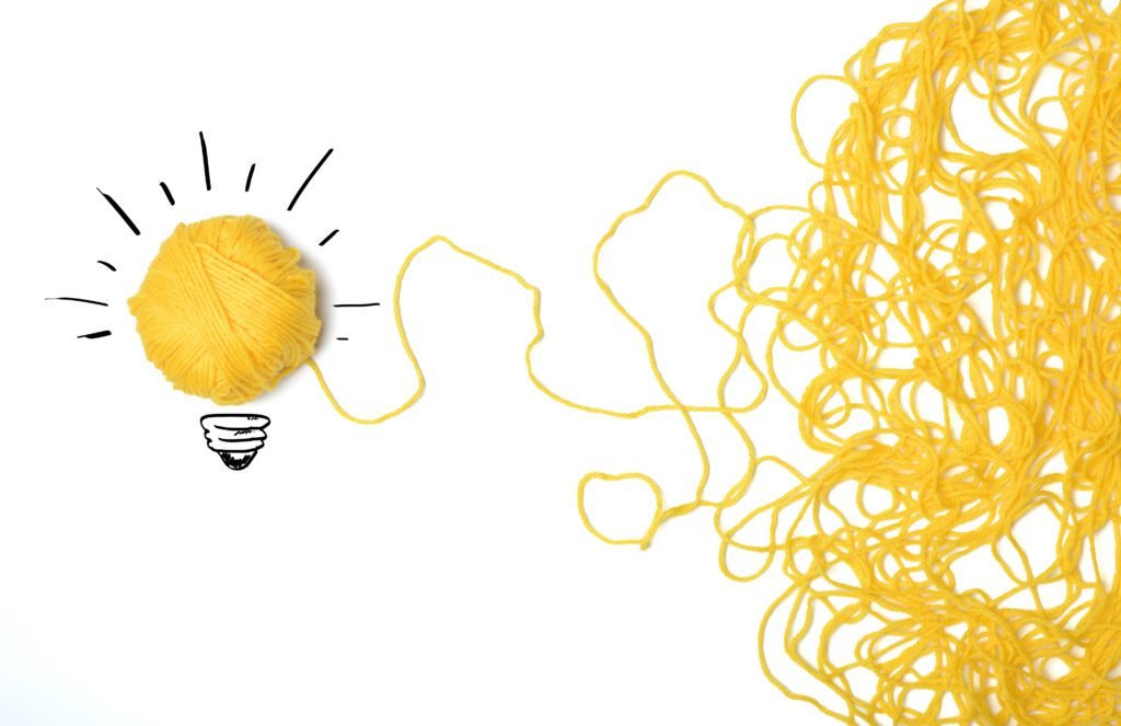 Descrição da imagem #PraCegoVer: Imagem no formato retangular, na horizontal. A imagem tem fundo branco. Do lado direito temos um novelo de lã amarela solto, que se transforma em uma lâmpada. Ao redor do novelo, que forma a lâmpada, temos o desenho de um bocal e traços circundando o novelo, remetendo à chama da lâmpada. Fim da descrição.