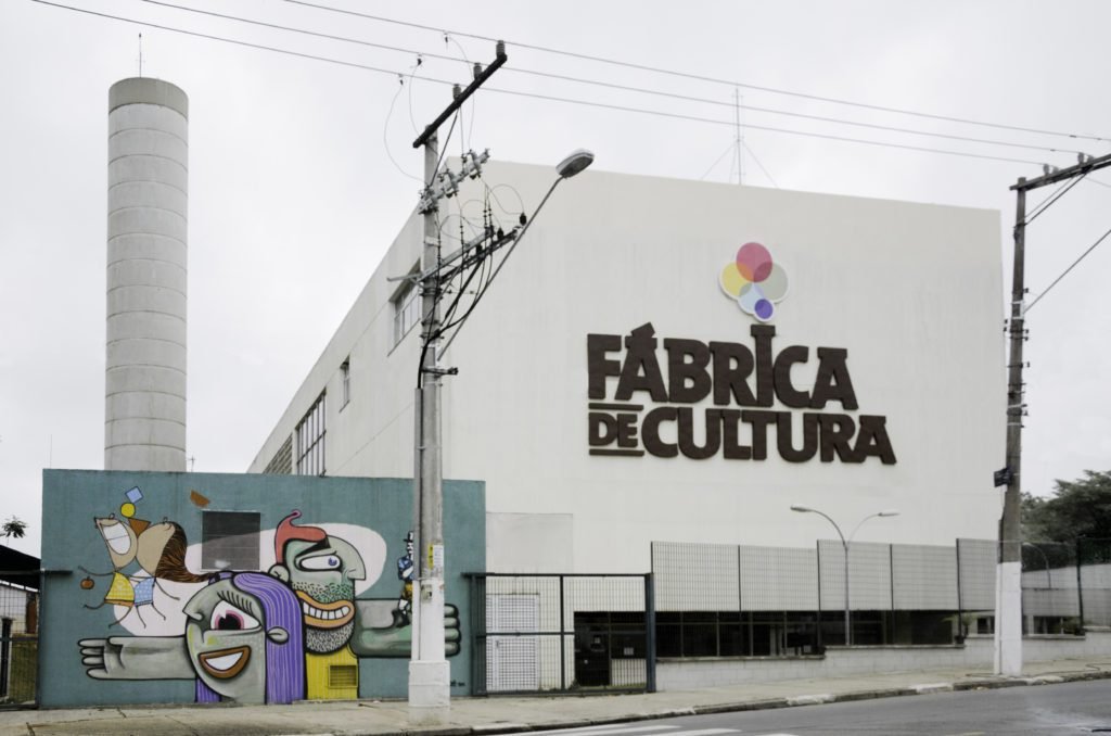 Descrição da imagem #PraCegoVer: Imagem no formato retangular, na horizontal. Fachada da Fábrica de Cultura do Jardim São Luís. Um prédio branco, com o nome: Fábrica de Cultura, escrito em letras pretas. No canto inferior, esquerdo, vemos um muro grafitado colorido, com predominância na cor verde. Fim da descrição.