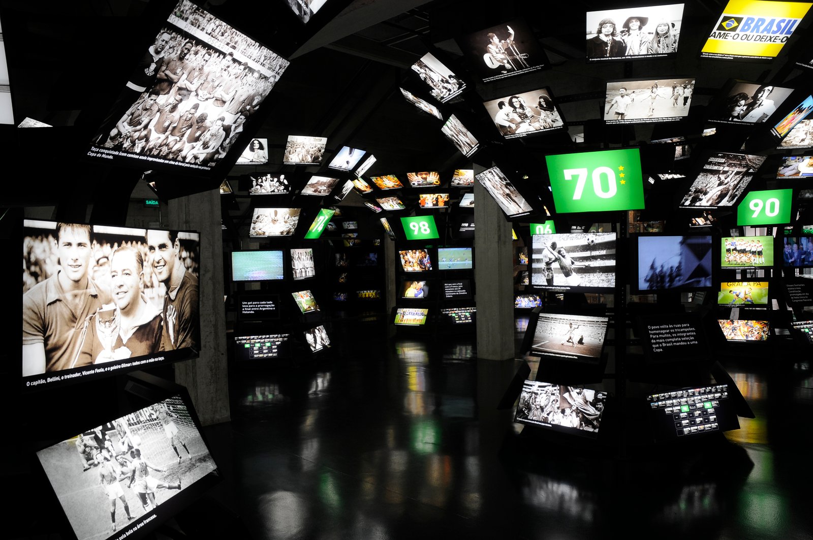 Descrição da imagem #PraCegoVer: Imagem no formato retangular, na horizontal. Uma das salas de exposição do Museu do Futebol. A sala é escura e tem diversas telas de led, com imagens de momentos históricos do futebol. Fim da descrição.