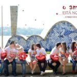Descrição da imagem #PraCegoVer: Imagem no formato retangular, na horizontal. Na imagem estão cinco casais de namorados, todos eles têm síndrome de Down. Eles estão sentados na frente da Igreja de São Francisco de Assis, na Pampulha. O local é considerado um cartão-postal da cidade de Belo Horizonte. Na parte superior da imagem foram inseridas imagens de pequenos corações e no canto superior direito está escrito na cor vermelha ‘O amor está no ar, e porque não estaria?’. Os casais estão vestindo camiseta branca e seguram balões vermelhos em formato de coração. Fim da descrição.