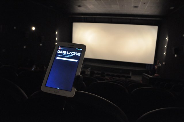 Descrição da imagem #PraCegoVer: Imagem no formato retangular, na horizontal. Uma sala de cinema, escura. Em primeiro plano vemos um tablet com o whatscine em uso. Ao fundo, temos branca a tela de projeção do cinema. Fim da descrição.
