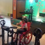 Descrição da imagem #PraCegoVer: Imagem no formato retangular, na horizontal. Sofia está tocando bateria, na área do museu em que as crianças são convidadas a formar uma banda musical Ao fundo, vemos uma outra criança tocando teclado. Fim da descrição.