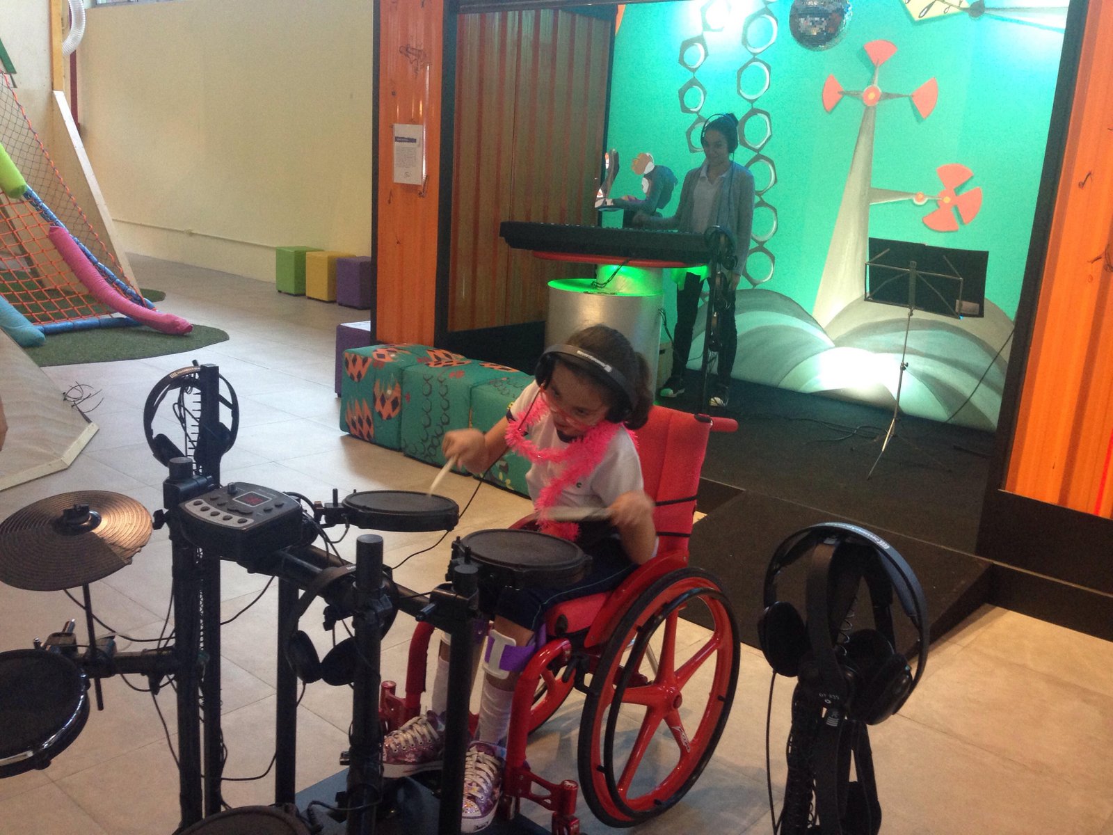 Descrição da imagem #PraCegoVer: Imagem no formato retangular, na horizontal. Sofia está tocando bateria, na área do museu em que as crianças são convidadas a formar uma banda musical Ao fundo, vemos uma outra criança tocando teclado. Fim da descrição.