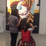 Descrição da imagem #PraCegoVer: Imagem no formato retangular, na vertical. Nela, Sofia está sentada em sua cadeira, tocando uma obra de arte do museu. Sofia está acompanhada de uma monitora do museu. Fim da descrição.