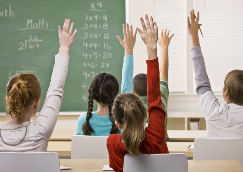 Descrição da imagem #PraCegoVer: Imagem no formato retangular, na horizontal. Nela, vemos cinco crianças sentadas em uma sala de aula. Vemos os alunos de costas, e à sua frente está a lousa, com algumas atividades de matemática. Todas as crianças estão com o braço levantado, como se tivessem solicitando a atenção do professor. Fim da descrição.