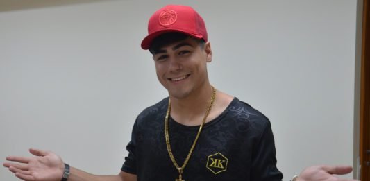 Descrição da imagem #PraCegoVer: Imagem no formato retangular, na horizontal. Nela, está o cantor Mc Milk. Ele é um rapaz jovem, com pele morena. Ele está com uma camiseta preta e usa um boné vermelho e um colar com um crucifixo. Ele está com os braços abertos e sorri. Fim da descrição.