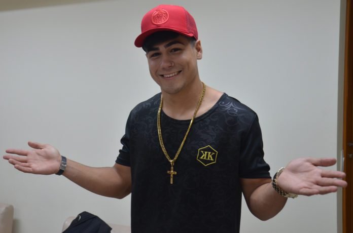 Descrição da imagem #PraCegoVer: Imagem no formato retangular, na horizontal. Nela, está o cantor Mc Milk. Ele é um rapaz jovem, com pele morena. Ele está com uma camiseta preta e usa um boné vermelho e um colar com um crucifixo. Ele está com os braços abertos e sorri. Fim da descrição.