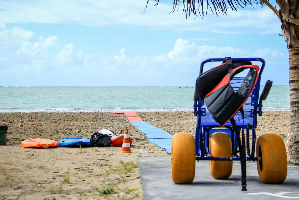 Descrição da imagem #pracegover: Foto de uma praia preparada para receber pessoas com deficiência. Na imagem vemos uma esteira na areia, que leva pessoas com deficiência física até o mar. Do lado esquerdo vemos alguns equipamentos de segurança e do lado direito, uma cadeira de rodas anfíbia. Ao fundo vemos o mar e no canto direito vemos um coqueiro. Fim da descrição.