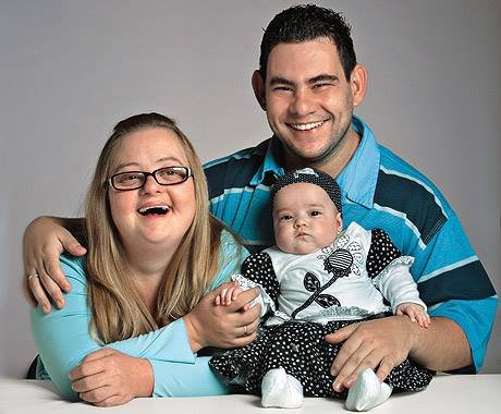 Descrição da imagem #PraCegoVer: Imagem no formato retangular, na horizontal. Nela estão Gabriela, que tem síndrome de Down, Fábio, que tem deficiência intelectual, e a pequena Valentina, filha do casal, que não tem deficiência. Eles estão sorrindo. Fim da descrição.
