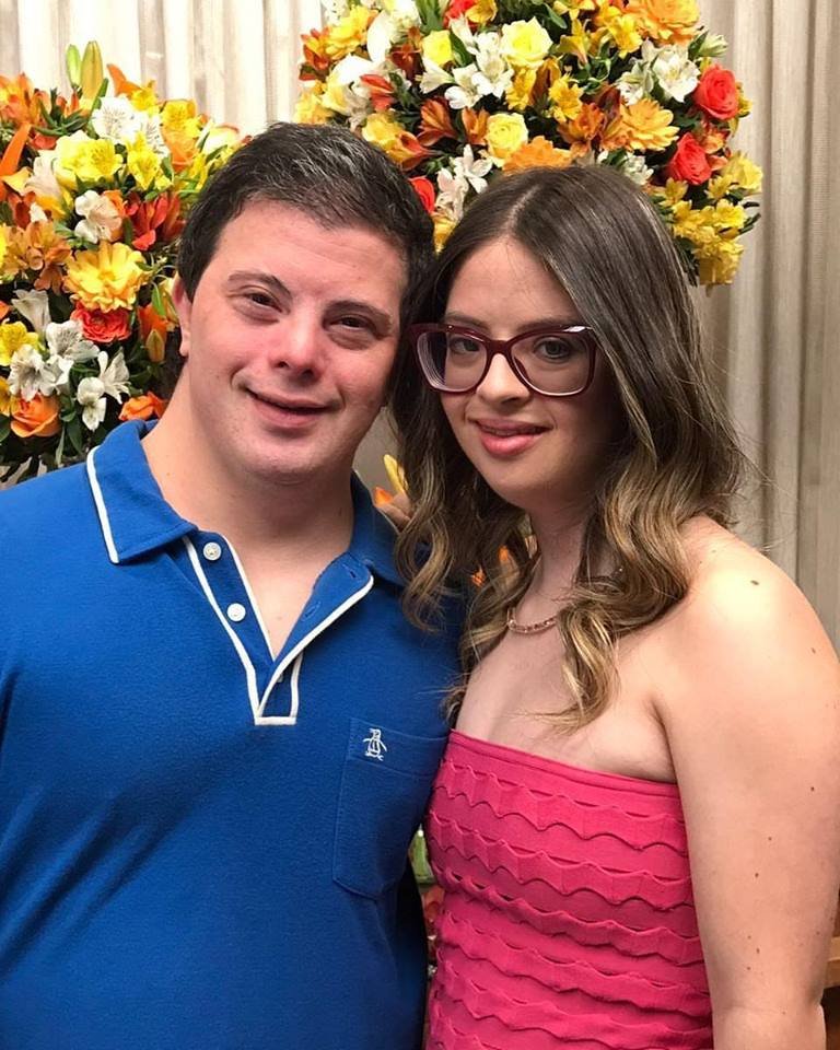 Descrição da imagem #PraCegoVer: Imagem no formato retangular, na vertical. Nela, o casal Breno e Samanta estão abraçados. Ele usa uma camiseta polo azul com detalhes em branco, e ela usa um vestido, tomara que caia, cor de rosa. Eles estão sorrindo. Ao fundo é possível ver um arranjo de flores coloridas. Fim da descrição.