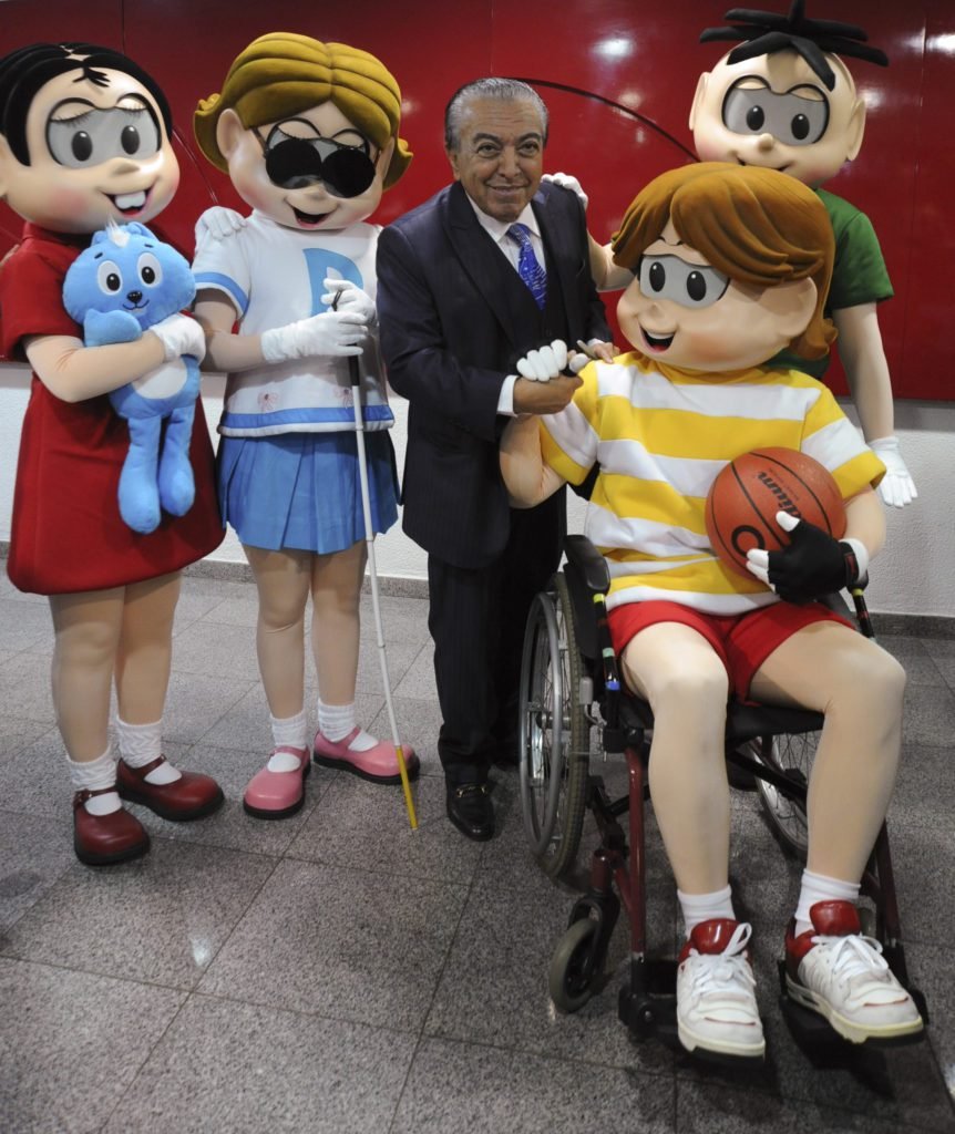 Descrição da imagem #PraCegoVer: Imagem no formato retangular, na vertical. Nela, o cartunista Maurício de Sousa está acompanhado dos personagens da Turma da Mônica. Na ordem, da esquerda para a direita, estão Mônica, Dorinha, que tem deficiência visual, Maurício de Sousa, Luca, que é cadeirante, e Cebolinha. Fim da descrição.
