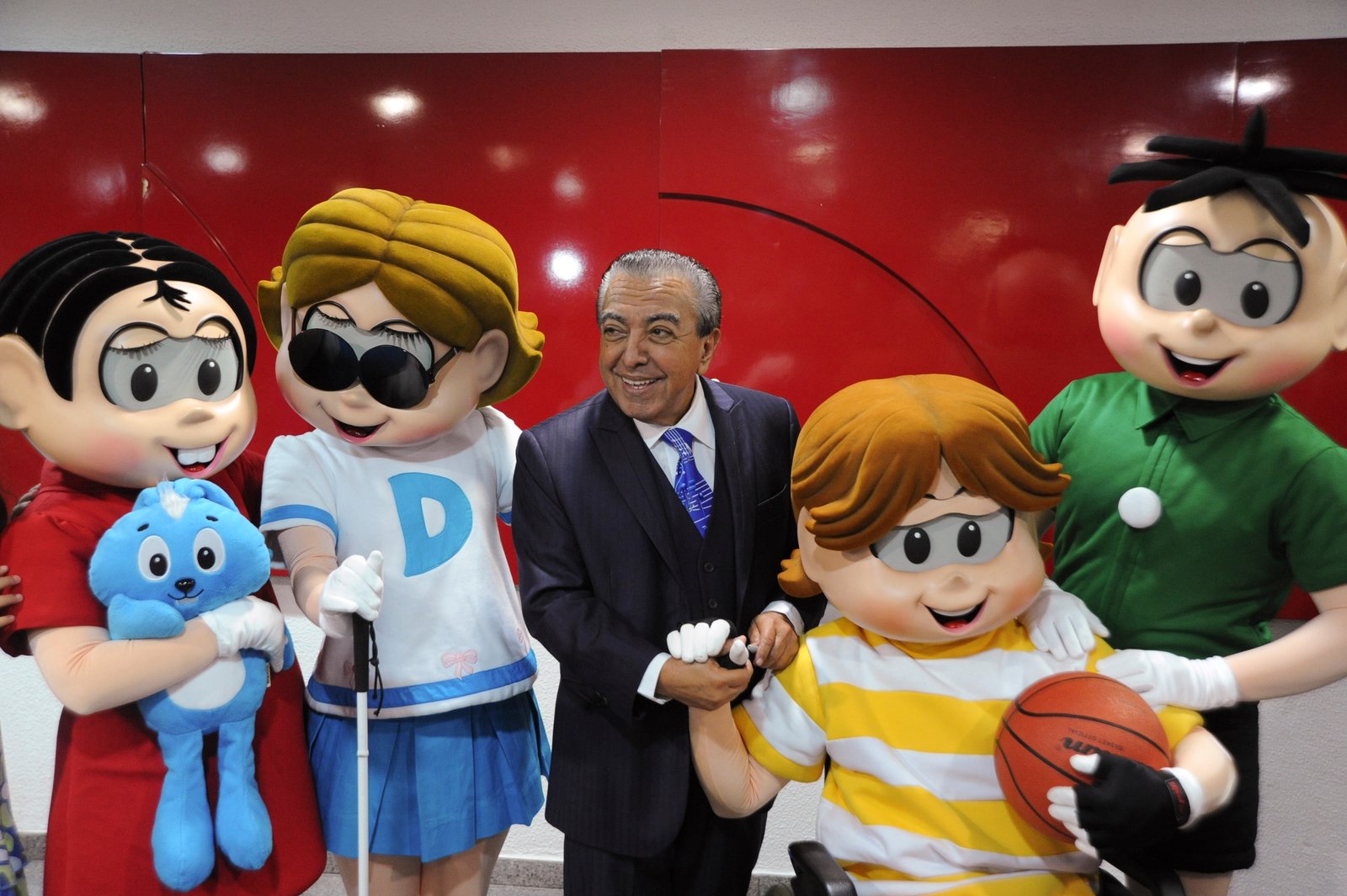 Descrição da imagem #PraCegoVer: Imagem no formato retangular, na horizontal. Nela, o cartunista Maurício de Sousa está acompanhado dos personagens da Turma da Mônica. Na ordem, da esquerda para a direita, estão Mônica, Dorinha, que tem deficiência visual, Maurício de Sousa, Luca, que é cadeirante, e Cebolinha. Fim da descrição.