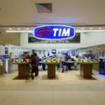 Descrição da imagem #PraCegoVer: A imagem está no formato retangular, na horizontal. A imagem tem a fachada de uma loja da TIM. É possível ver aparelhos de celular e tablets expostos em balcões de madeira. Acima está o logo da empresa, que consiste em um painel na cor azul com três traços ondulados em vermelho e o nome TIM em letras maiúsculas, em branco. Fim da descrição.