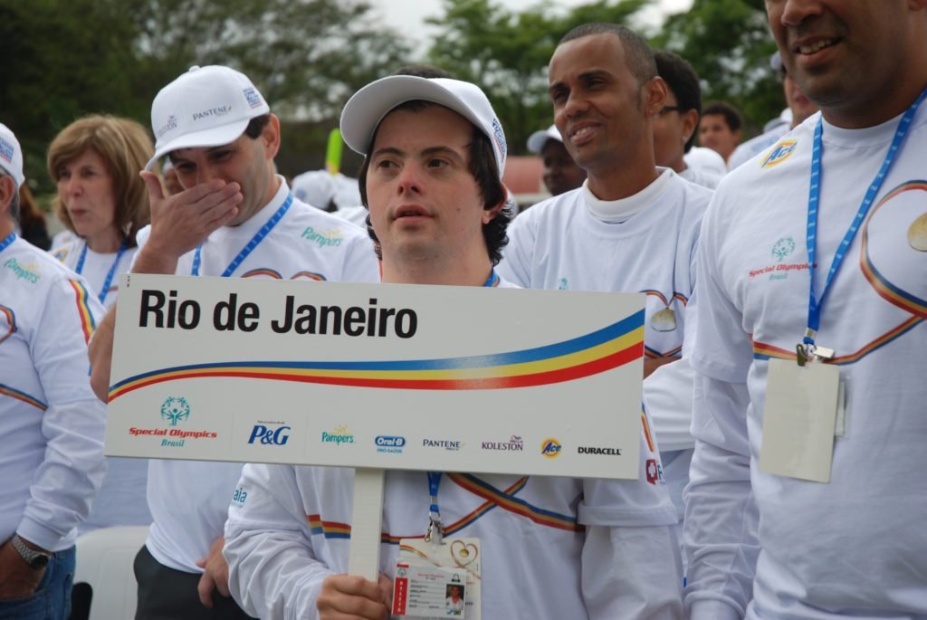 Descrição da imagem #PraCegoVer: Imagem no formato retangular, na horizontal. Nela está o atleta do judô Breno Viola, que representou o Brasil em diversos eventos da Special Olympics. Ele está entre outros atletas da entidade, durante uma cerimônia de abertura de evento. Ela usa uniforme e boné branco, e segura uma placa onde está escrito: Rio de Janeiro, Special Olympics Brasil. A placa também traz alguns logos de patrocinadores. Fim da descrição.