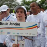 Descrição da imagem #PraCegoVer: Imagem no formato retangular, na horizontal. Nela está o atleta do judô Breno Viola, que representou o Brasil em diversos eventos da Special Olympics. Ele está entre outros atletas da entidade, durante uma cerimônia de abertura de evento. Ela usa uniforme e boné branco, e segura uma placa onde está escrito: Rio de Janeiro, Special Olympics Brasil. A placa também traz alguns logos de patrocinadores. Fim da descrição.
