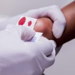 Descrição da imagem #PraCegoVer: A imagem está no formato quadrado. Nela é possível ver a realização do Teste do Pezinho. Uma profissional da área de saúde faz a coleta do material no pé de um bebê recém-nascido. Na imagem é possível ver a placa de coleta marcada pelo sangue da criança. Fim da descrição.