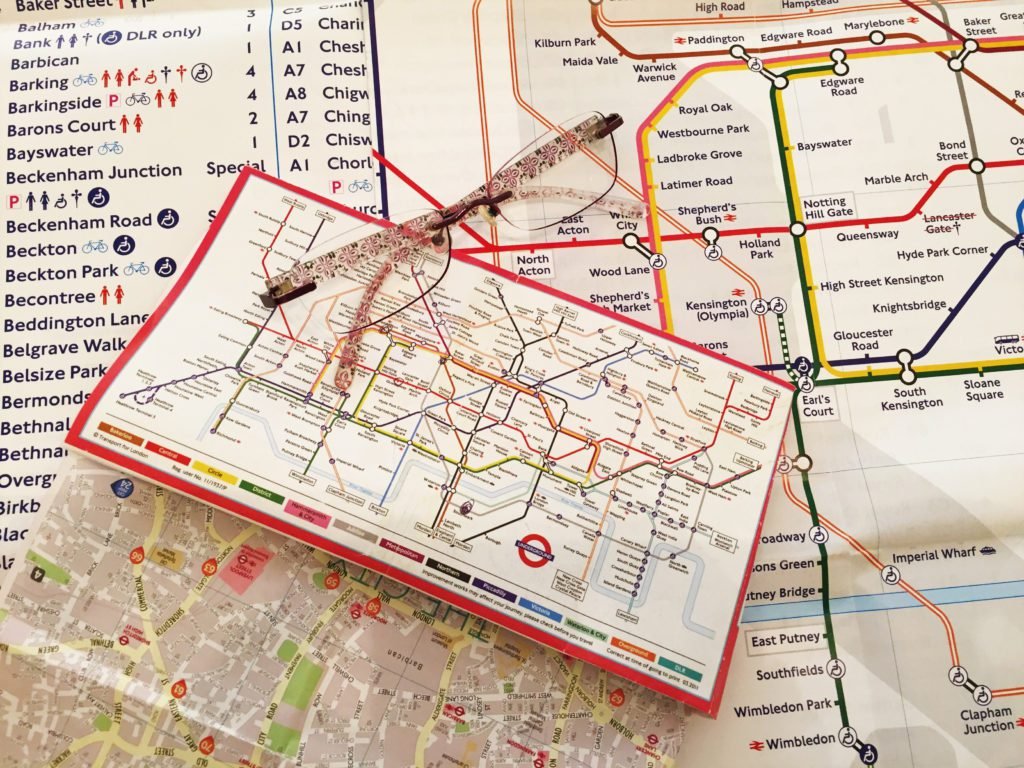 Descrição da imagem #PraCegoVer: A imagem está no formato retangular, na horizontal. Nela estão dois mapas do metrô de Londres. Um com letras grandes aberto em cima de uma mesa e outro com letras muito pequenas, dobrado, em cima do outro mapa, que está aberto. Sobre eles está um óculos de leitura. Fim da descrição.