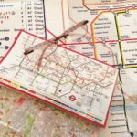 Descrição da imagem #PraCegoVer: A imagem está no formato retangular, na horizontal. Nela estão dois mapas do metrô de Londres. Um com letras grandes aberto em cima de uma mesa e outro com letras muito pequenas, dobrado, em cima do outro mapa, que está aberto. Sobre eles está um óculos de leitura. Fim da descrição.