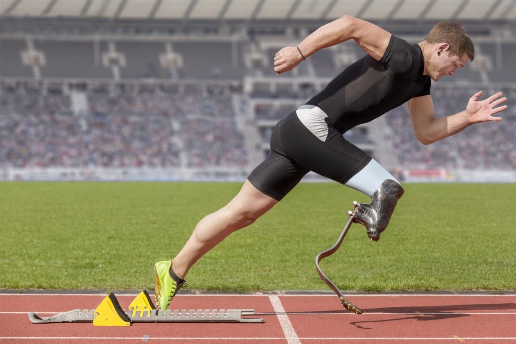 Descrição da imagem #PraCegoVer: A imagem está no formato retangular, na horizontal. Ela tem em destaque um paratleta de atletismo em um estádio. Ele está de lado, em posição de largada. O atleta usa uma prótese especial para corrida na perna direita. Ao fundo é possível ver a arquibancada do estádio cheia de pessoas. Fim da descrição.