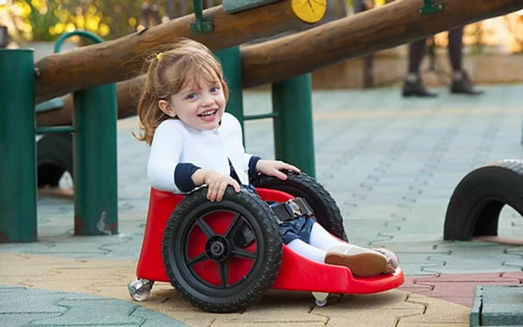 Descrição da imagem #PraCegoVer: A imagem está no formato retangular, na horizontal. Nela está a pequena Ana Paula em seu carrinho Fly Children. O carrinho é vermelho, tem duas rodas pretas e Ana está usando um vestido azul, estampado, uma blusa e meias branca. Ela está em um parque de diversões e sorri. Fim da descrição.