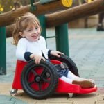 Descrição da imagem #PraCegoVer: A imagem está no formato retangular, na horizontal. Nela está a pequena Ana Paula em seu carrinho Fly Children. O carrinho é vermelho, tem duas rodas pretas e Ana está usando um vestido azul, estampado, uma blusa e meias branca. Ela está em um parque de diversões e sorri. Fim da descrição.